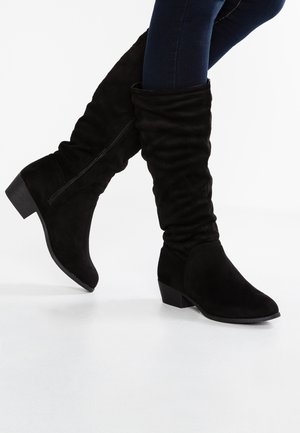 Zwarte suède laarzen tot halverwege de kuit met lage blokhakken, gedragen bij donkerblauwe skinny jeans op een witte achtergrond.