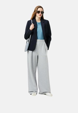 Femme portant un pantalon de survêtement gris oversize, une chemise bleue, un blazer noir, des baskets blanches, des lunettes de soleil et portant un sac à épaule gris, debout devant un fond blanc.