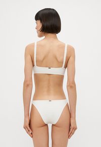 Balts bikini komplekts, kas sastāv no teksturēta braleta ar volāna siksniņām un atbilstošiem hipster stila apakšbiksēm, vienkārša dizaina, bezšuvju apdare.