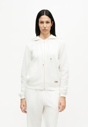 Femme aux cheveux lisses et foncés portant un sweat à capuche blanc zippé et un pantalon blanc assorti, debout devant un fond blanc uni.