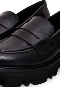 Mocassins en cuir noir avec des semelles épaisses et robustes à crampons, et un détail lisse de bride penny sur la partie avant supérieure.