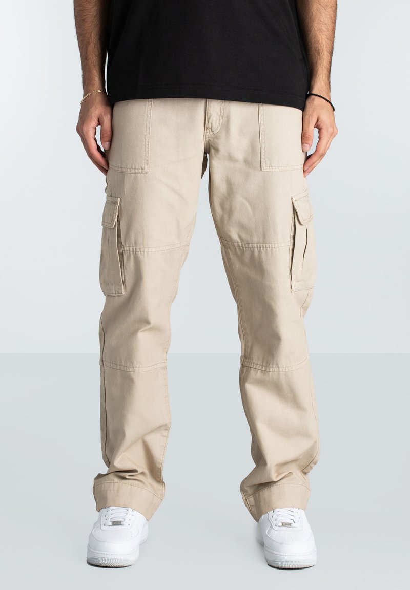 Beige cargo pants with side pockets, straight legs, and slight tapering at the ankles, paired with white sneakers and a black t-shirt.