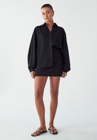 Černá košile na knoflíky s balónovými rukávy, spárovaná s černou mini sukní s vysokým pasem. Model má na sobě černé ploché sandály. Minimalistický design.