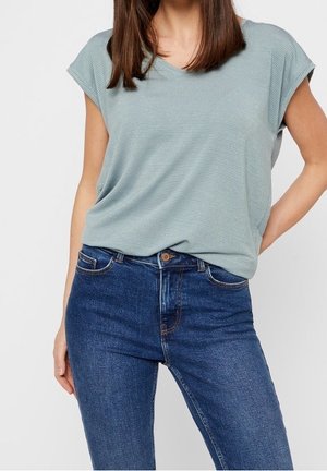 Kobieta w jasnoniebieskim, prążkowanym, krótkim topie włożonym w ciemnoniebieskie, wysokowentylowane dżinsy, pozująca z jedną ręką na biodrze.