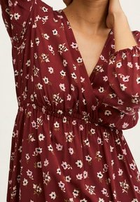 Tmavě červené květinové šaty s výstřihem do V a elastickým pasem. Mají nabírané rukávy a malé bílé a růžové květinové vzory po celé látce.