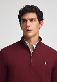 Sudadera de cuello con cremallera en burdeos, confeccionada en un material de punto suave, con textura acanalada sutil y un pequeño logotipo bordado en el pecho.