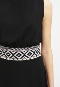 Vestido negro sin mangas con un cinturón ancho tejido que presenta patrones geométricos en blanco y negro en forma de diamante alrededor de la cintura.