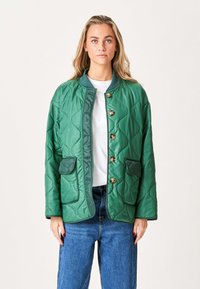 Quilted grøn jakke med rund krave, der har to lommer, guldbuttons og en glat tekstur. Kombineret med en hvid skjorte og jeans.