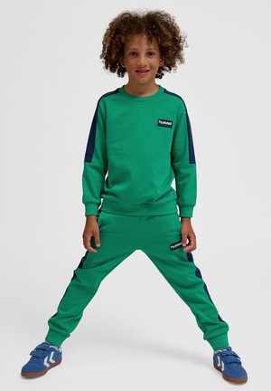 Grünes Sweatshirt- und Hosen-Set mit marineblauen Seiteneinsätzen, Rippbündchen und einem Logo-Patch. Die Schuhe sind blau mit Klettverschlüssen und braunen Sohlen.