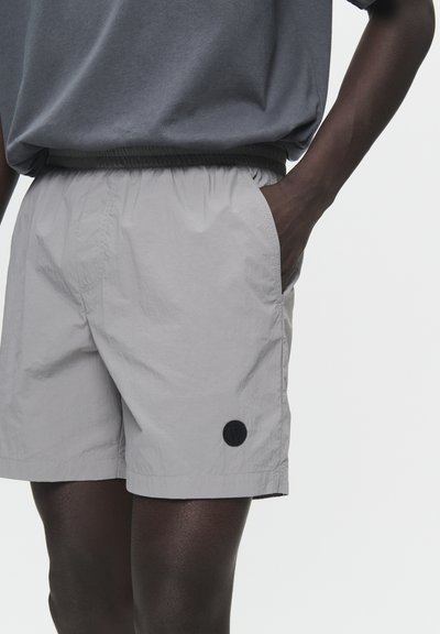 Lys grå shorts lavet af letvægtsstof, med elastisk talje og sidelommer; inkluderer en sort rund logo-patch.