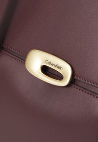 Sac à main bordeaux en matériau synthétique lisse, avec quincaillerie dorée à ton et un détail découpé, et des accents de couture contrastés.