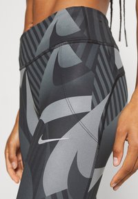 Zbliżenie osoby noszącej czarno-szare legginsy w wzór od Nike, z widocznym szwem bocznym i małym białym logo Nike swoosh.
