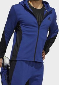 Veste zippée bleue avec des accents noirs, tissu texturé et capuche. Fait partie d'un ensemble assorti avec un t-shirt et un pantalon de sport noirs.