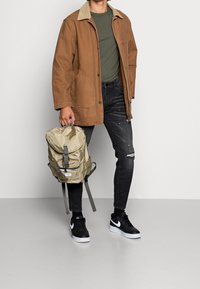 Chaqueta de lona marrón con cuello beige, camisa verde oliva, vaqueros negros desgastados, zapatillas negras y blancas, y mochila beige.