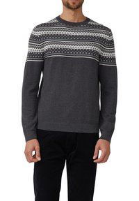 Homme portant un pull gris foncé avec des motifs géométriques blancs et gris clair sur la poitrine et les manches supérieures, associé à un pantalon noir.