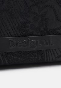Schwarze Lederhülle mit strukturierten Mustern und gestickten Akzenten, versehen mit einem hervorgehobenen Logo-Band mit der Aufschrift "Desigual". Insgesamt glatte Oberfläche.