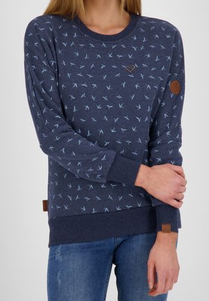 Femme portant un pull bleu marine avec de petits motifs d'oiseaux, un patch cœur sur la poitrine et un jean bleu. Mains croisées à la taille.
