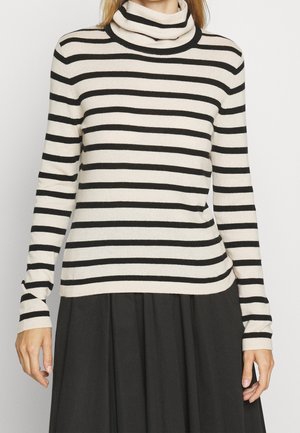 Femme portant un pull à col roulé beige à manches longues avec des rayures horizontales noires et une jupe plissée noire, devant un fond uni.