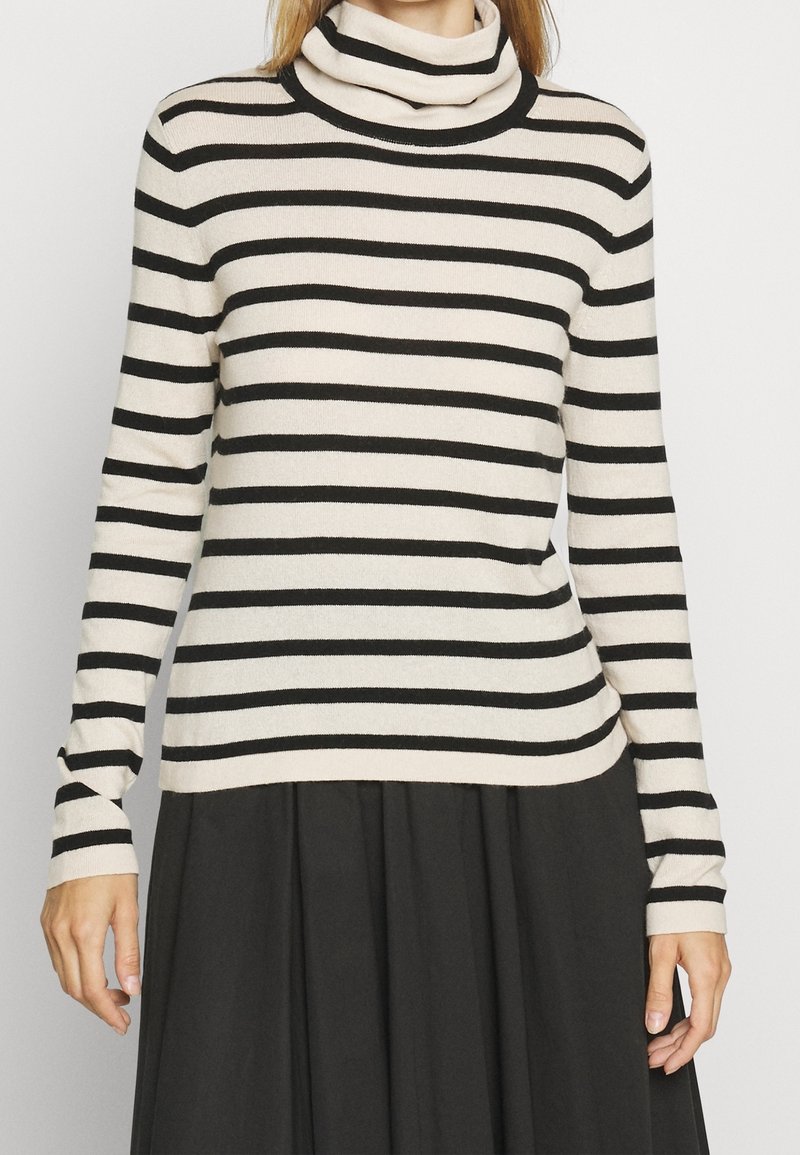 Femme portant un pull à col roulé beige à manches longues avec des rayures horizontales noires et une jupe plissée noire, devant un fond uni.