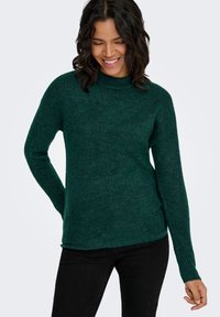 Dunkelgrüner Strickpullover mit hohem Kragen und langen Ärmeln, der eine strukturierte Oberfläche und seitliche Schlitze am Saum aufweist. Darunter schwarze Hosen.