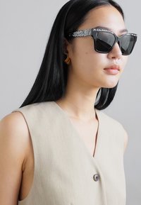 Svarta oversized solglasögon med glittrande detaljer på bågarna. Modellen bär en beige, ärmlös topp med knäppning.
