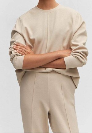 Osoba w beżowej długiej bluzce z rękawami i dopasowanych spodniach stoi ze skrzyżowanymi ramionami na jednolitym tle.