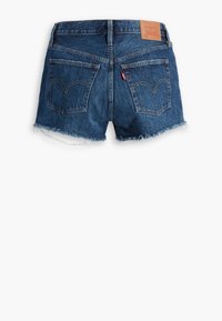 Short en denim bleu avec ourlets effilochés, deux poches arrière, une petite étiquette rouge sur la poche droite et un patch en cuir avec le logo de la marque sur la ceinture.