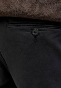 Povečan posnetek zadnjega žepa črnih hlač z gumbom in temno rjavim robom pletenega puloverja nad njim.