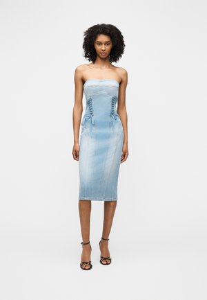 Nainen pukeutuneena vaaleansiniseen olkahihnattomaan farkkumekkoon ja mustiin korkeakorkoisiin sandaaleihin, seisomassa yksinkertaista vaaleanharmaata taustaa vasten.