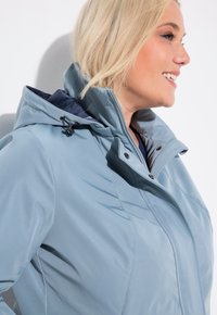 Ljusblå vattentät jacka med hög krage, dragkedjestängning och justerbara snoddar. Mjuk textur med söm-detaljer och tryckknappar.