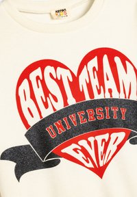 Sudadera crema con un atrevido diseño de corazón rojo que incluye el texto "MEJOR EQUIPO DE LA HISTORIA" y una banda negra con la etiqueta "UNIVERSIDAD", adornada con destellos.