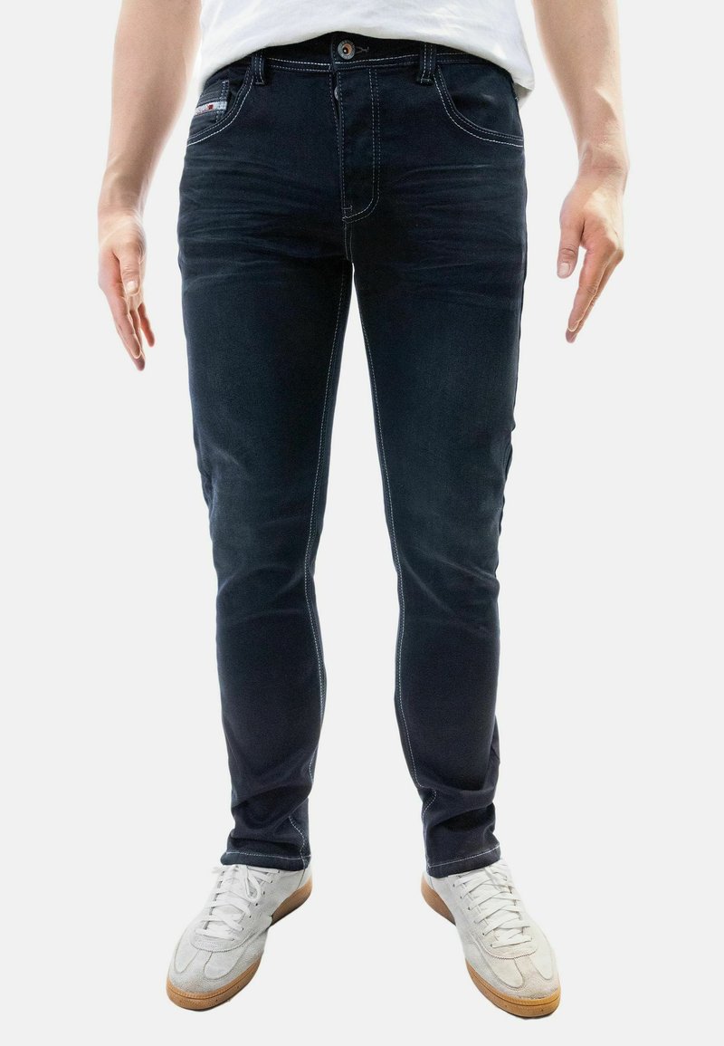 Dark blue denim jeans with a slim fit, light contrast stitching, five pockets, and a button closure, worn with white athletic shoes.