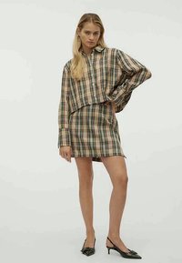 Sieviete, tērpusies bēšas krāsas iekšējā pārklājumā ar rūtiņām, valkā piegulošu svārku un pozē ar roku uz gurniem, melnās šaurās kurpēs ar atvērtu papēdi.