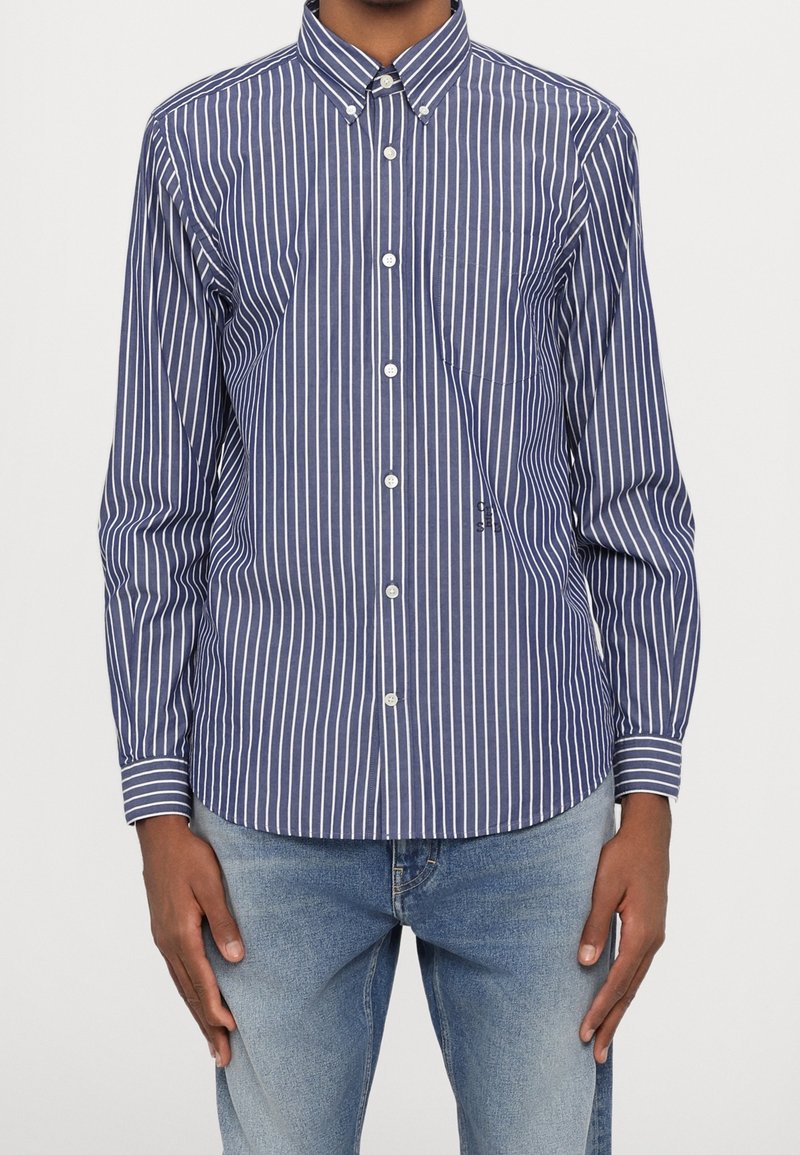 Homme portant une chemise boutonnée à rayures verticales bleu et blanc avec col et un jean bleu clair, debout avec les bras détendus le long du corps.