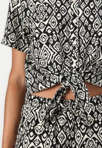 Chemise courte à motifs noir et blanc avec des manches courtes, nouée à la taille, présentant un design géométrique et des boutons sur le devant.
