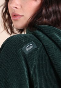 Veste en velours côtelé vert foncé avec une capuche et une étiquette de marque "OXBOW" bien visible sur l'épaule. Tissu texturé avec un motif côtelé.