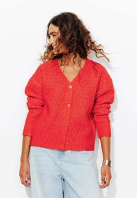 Cardigan tricoté rouge avec un motif en diamant, en col en V et avec des poignets côtelés. Caractérisé par de gros boutons et une coupe décontractée. Associé à un jean bleu clair.