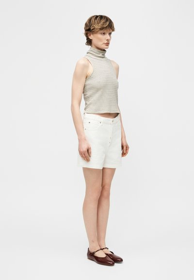 Γυναίκα που στέκεται πλάγια, φοράει αμάνικη ριγέ μπλούζα με ζιβάγκο, λευκά σορτς ψηλής μέσης και σκούρα καφέ παπούτσια Mary Jane.