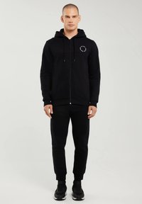 Bărbat stând cu fața spre înainte, purtând hanorac negru cu fermoar, pantaloni sport și adidași negri, pe un fundal simplu, luminos.