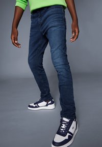 Ciemnoniebieskie jeansy slim-fit wykonane z denimu, z detalicznym szyciem wzdłuż szwów, zestawione z białymi i granatowymi trampkami.