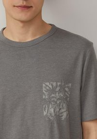 Grijze katoenen T-shirt met een ronde hals. Voorzien van een gestreepte zak met palmbladeren in een lichtere grijze tint, wat textuur en detail toevoegt.