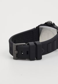 Black silicone watch strap with a textured surface and metal buckle. Features include embossed branding and adjustable holes for fitting.
