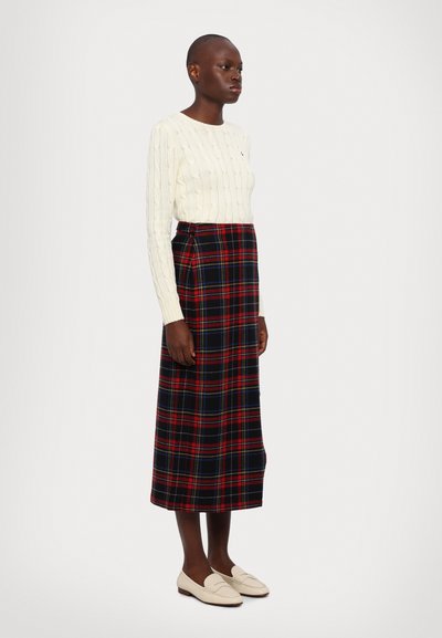 Maglione di lana a trecce crema, gonna lunga a quadri in tonalità rosse, nere e blu, e mocassini bianchi. Tessuto strutturato con silhouette dritta.