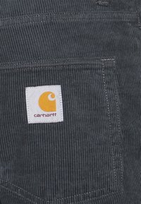 Donkergrijze ribfluweel stof met een vierkant wit Carhartt-logo als patch, vastgenaaid op een zak.