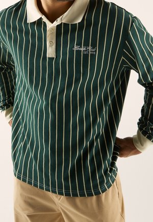 Mann trägt dunkelgrünes langärmeliges Poloshirt mit cremefarbenen vertikalen Streifen, cremefarbenem Kragen und Manschetten und Logo "Health Club EST. 1998".