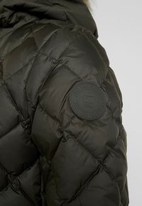 Gros plan sur l’épaule d’une veste matelassée sombre avec un patch en caoutchouc circulaire indiquant "Just the product" et le logo "RAW".