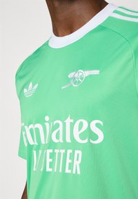 Grünes Sporttrikot mit weißen Akzenten, mit dem Schriftzug "Emirates Fly Better" und einem Logo, das eine Kanone zeigt. Hergestellt aus leichtem, glattem Stoff.