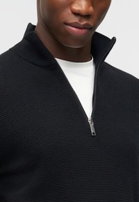 Άνδρας που φοράει μαύρο ριμπ μισό φερμουάρ πουλόβερ πάνω από λευκό μπλουζάκι με στρογγυλή λαιμόκοψη, το φερμουάρ είναι μερικώς ανοιχτό, πάνω σε απλό φόντο.