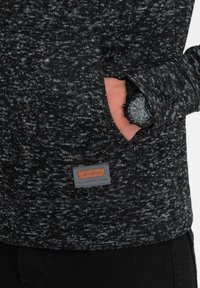 Zwarte en grijze gestructureerde sweatshirt met een voorzak, voorzien van een gestikt label, gedragen over donkere broek. Een zwart horloge is zichtbaar om de pols.