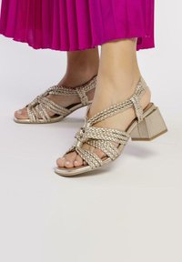Sandalias de oro trenzadas con tacón cuadrado, que presentan correas trenzadas y un diseño de punta abierta. El acabado texturizado complementa el aspecto general.